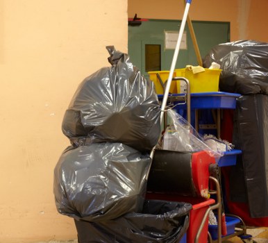 The image shows a collection of black rubbish bags filled with waste, stacked on a blue trolley and a red dolly, situated against a yellow wall in an indoor or enclosed space. The bags are made of shiny plastic and appear tightly packed, with some of the bags leaning against each other. A yellow dustpan and mop are positioned behind the refuse, suggestive of cleaning supplies. The background features a green door with a small window and a few shelves or containers, possibly used for storing cleaning or waste management tools. The scene is well-lit, emphasizing the textures of the plastic bags and the surfaces of the trolley, which is used for transporting rubbish. This setup is indicative of a waste collection or disposal area, potentially within a commercial or residential building, aligned with services provided by Business Waste Removal Dalston in the area postcode, supporting local rubbish removal needs.