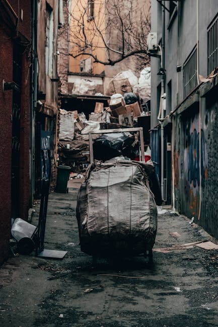 A narrow urban back alleyway filled with a large, grey, plastic rubbish bag on a wheeled trolley in the foreground, with dark, asphalt ground showing signs of dirt and debris. Behind the trolley, ther