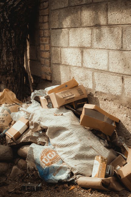 A narrow urban back alleyway filled with a large, grey, plastic rubbish bag on a wheeled trolley in the foreground, with dark, asphalt ground showing signs of dirt and debris. Behind the trolley, ther