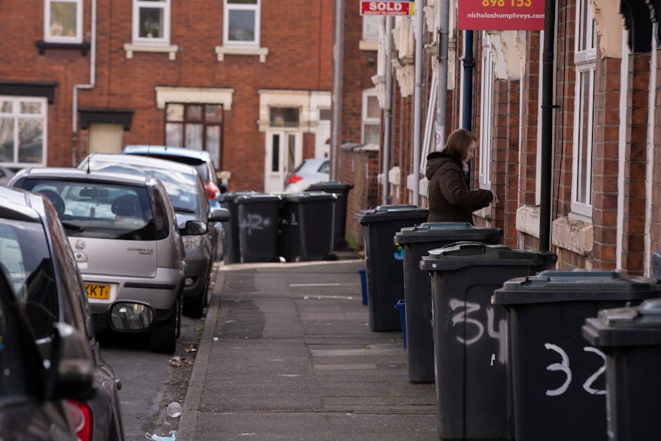 A black plastic wheelie bin with a white label and the text 'ST. JOHN'S' printed on it, positioned on a dimly lit urban sidewalk at night. The bin contains a mix of waste materials, including flattene